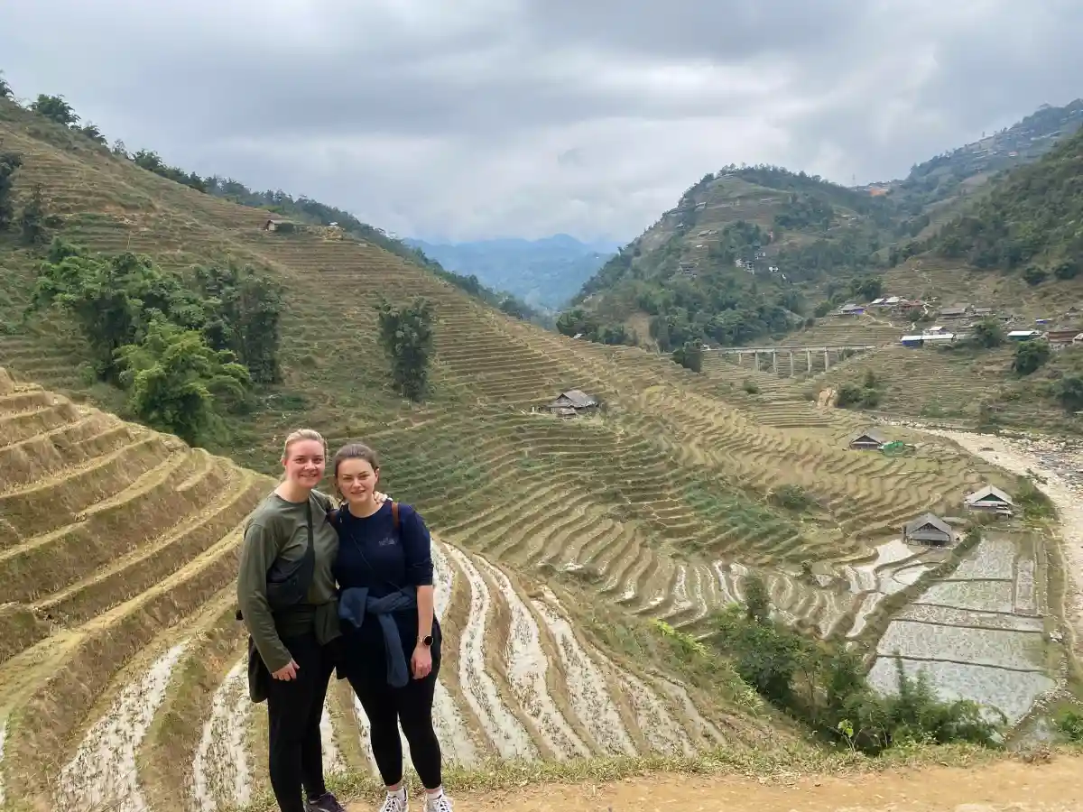 Two travellers on the 2-day Sapa homestay trek standing in front of layered rice terraces and a village