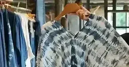 Smiling Hmong artisan holding up a handmade indigo tie-dye shirt inside a Sapa craft shop filled with batik fabrics and indigo garments