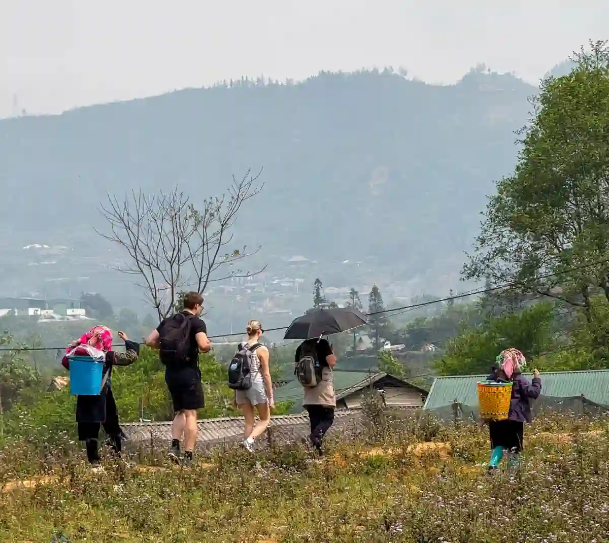 Trekkers walking with H'mong women carrying baskets through grass fields above Sapa town — typical arrival scene after overnight travel from Hanoi